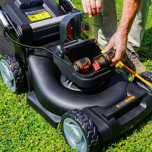 Worx 40V brushless self-propelled lawn mower with 46cm cutting width displayed on grass, showing open battery compartment with two orange and black lithium-ion batteries being inserted, black mower deck with gray wheels, and control panel visible in background