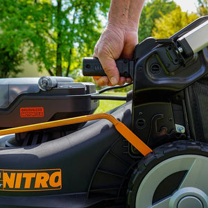 Close-up of a man's hand gripping the black handle of a Worx 40V brushless self-propelled lawn mower with orange and black trim, showing the motor label and wheel detail in an outdoor garden setting with green trees in the background