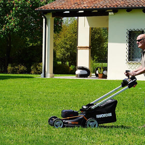 Man using Worx 40V Brushless Self Propelled Battery Lawn Mower with 46cm cutting deck on green lawn in front of modern white house with terracotta roof and black pergola