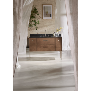 Mid-century modern bathroom vanity unit in light oak wood with black countertop, chrome handles and legs, featuring two drawers and cabinet doors, displayed in a minimalist bathroom with cream brick walls, framed abstract artwork, potted plant, and sheer white curtains