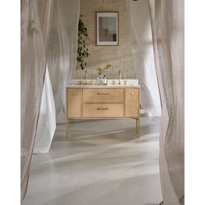 Light oak vanity unit with white marble countertop and brass fittings in modern bathroom with sheer white curtains and brick wall backdrop