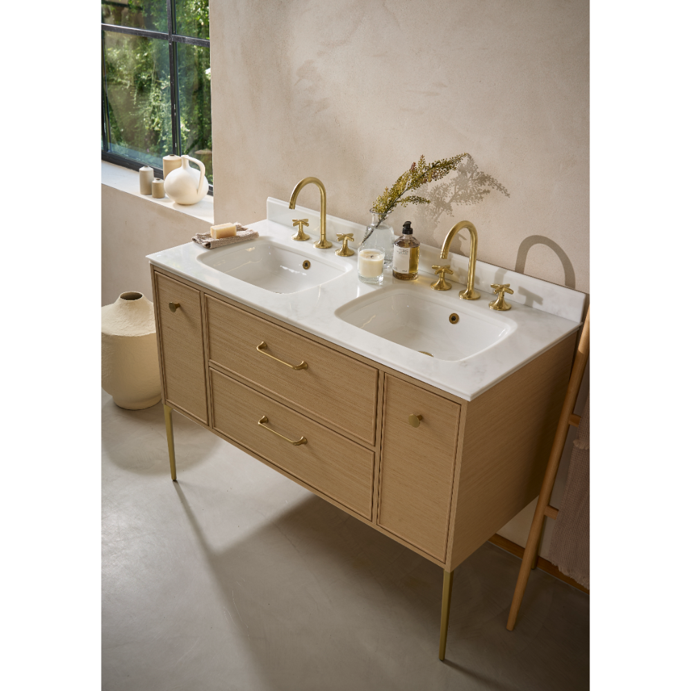 Double basin bathroom vanity unit in light oak wood with white marble countertop, brass faucets and handles, featuring two rectangular ceramic sinks with decorative accessories and potted plant
