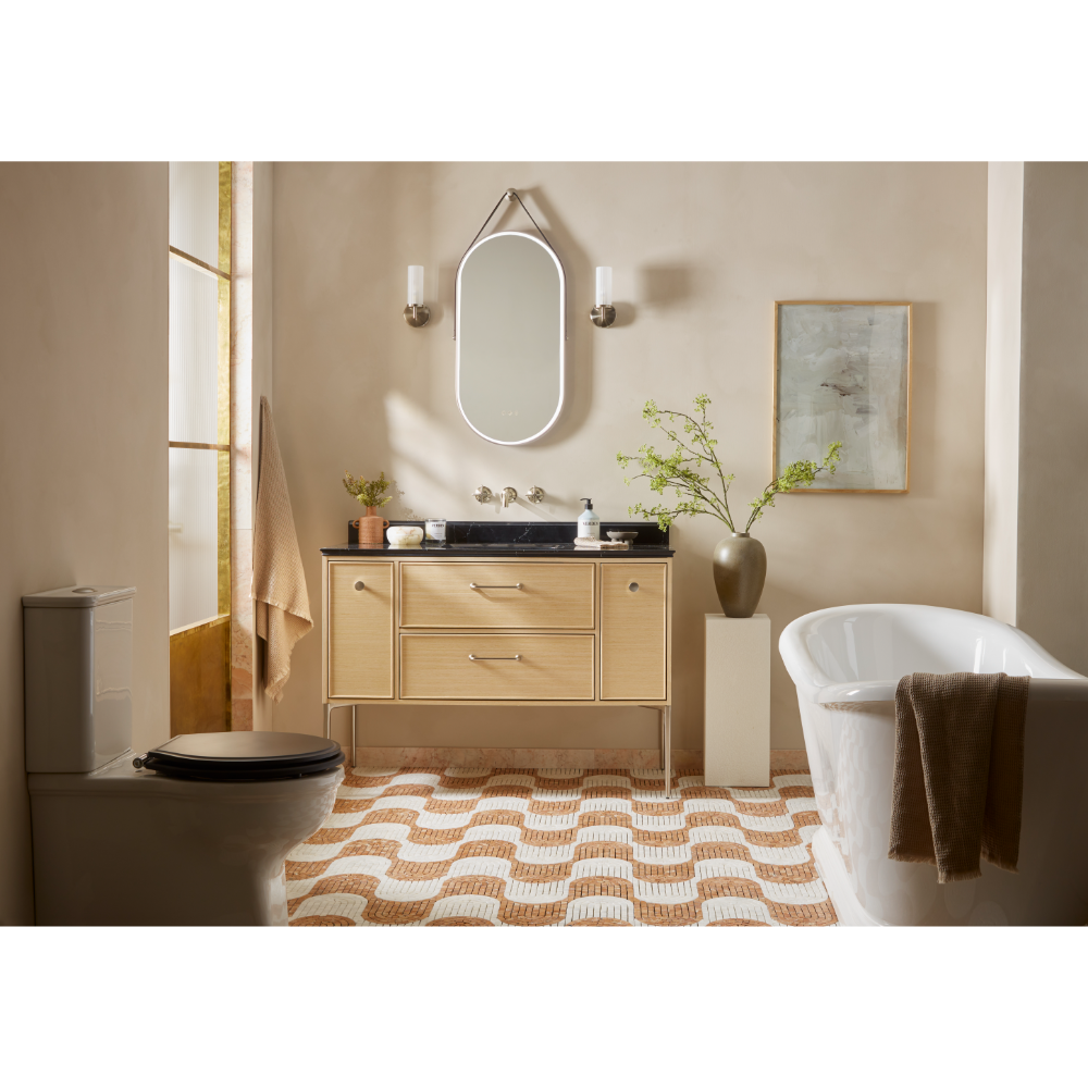 Light oak bathroom vanity unit with black marble countertop and integrated sink displayed in a warm beige bathroom with freestanding white bathtub, oval illuminated mirror, geometric patterned floor tiles, and decorative accessories
