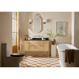 Light oak bathroom vanity unit with black marble countertop and integrated sink displayed in a warm beige bathroom with freestanding white bathtub, oval illuminated mirror, geometric patterned floor tiles, and decorative accessories