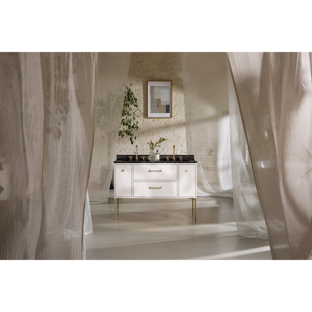 Modern white vanity unit with black countertop and brass handles in minimalist bathroom with exposed brick wall, framed mirror, potted plant, and sheer curtains