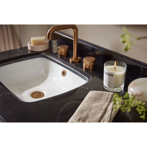 Modern bathroom vanity with white ceramic curved sink, black marble countertop, brushed bronze tap and faucet handles, soap dish, scented candle, gray towel and green plant decoration