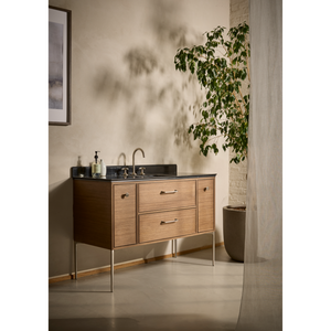 Light oak bathroom vanity unit with dark countertop and bronze fixtures, featuring two drawers and cabinet storage, set against white wall with climbing ivy plant and concrete column detail