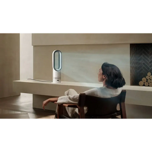 Woman sitting at desk facing white and silver Dyson Hot + Cool HF1 heater fan in minimalist beige interior with wooden chair and shelving