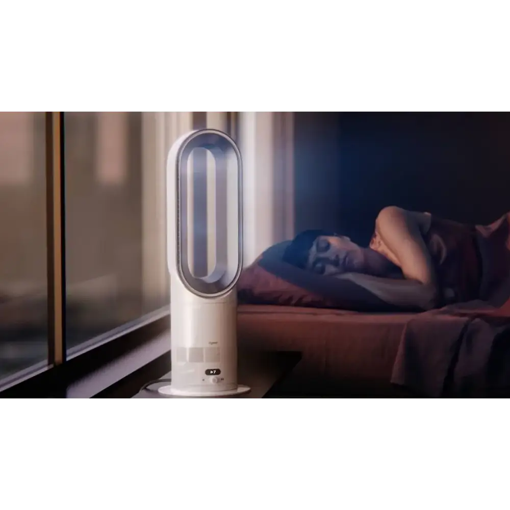 White and silver Dyson Hot + Cool HF1 heater and fan with oval-shaped design displayed on a nightstand next to a bed with a sleeping person in a modern bedroom setting