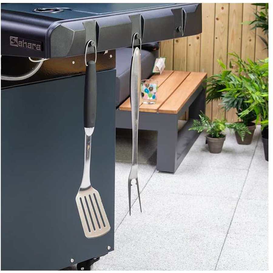 Black Sahara X450-S four burner gas BBQ grill with stainless steel utensils hanging from rail, wooden side table, potted plants and patio setting