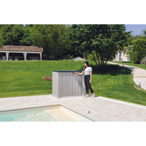 Woman opening a taupe grey and anthracite Toomax Storer Plus garden storage unit on a patio beside a swimming pool, with manicured lawn and residential buildings in the background