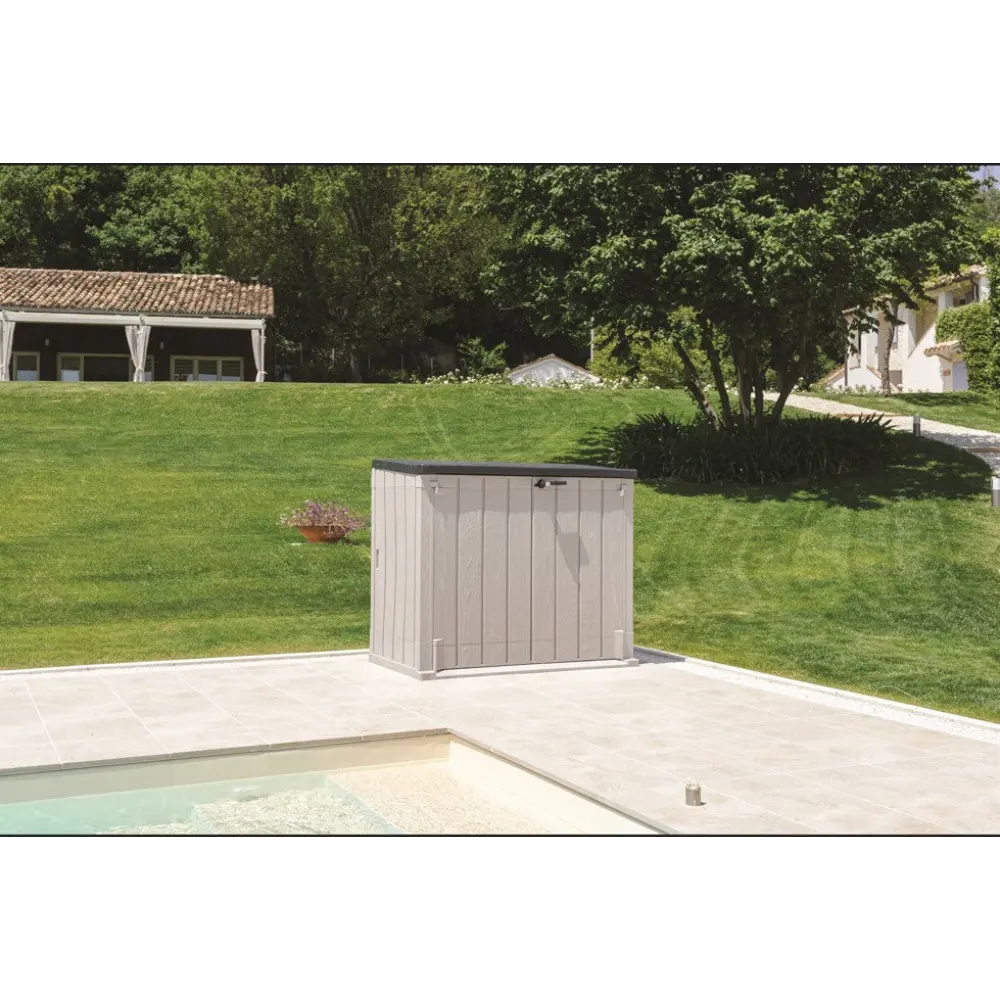 Toomax Storer Plus taupe grey and anthracite garden storage unit positioned on a poolside patio with manicured lawn, trees and residential buildings in background