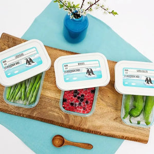 Three Keeeper food storage containers with turquoise lids arranged on a wooden cutting board atop a light blue surface, containing green beans, mixed berries, and snap peas, with a blue vase of white flowers and wooden spoon visible, displaying product labeling on white lids