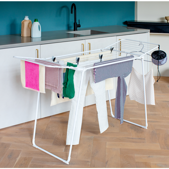 White metal foldable clothes drying rack with multiple horizontal bars holding various colorful garments including pink towel, green cloth and striped shirts in a modern kitchen with teal backsplash and white cabinetry