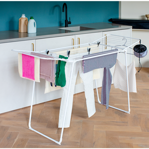 White metal foldable clothes drying rack with multiple horizontal bars holding various colorful garments including pink towel, green cloth and striped shirts in a modern kitchen with teal backsplash and white cabinetry