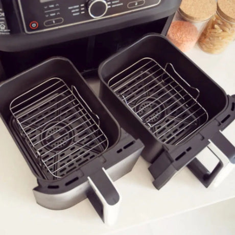 Two stainless steel wire basket inserts for air fryers with black non-stick coating, displayed next to an air fryer unit with control panel and condiment bottles on white surface