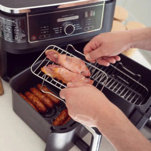 Two hands inserting stainless steel wire shelves into a black air fryer basket containing cooked chicken pieces and sausages, demonstrating the stackable cooking capacity of the Kitchen Academics air fryer accessory set