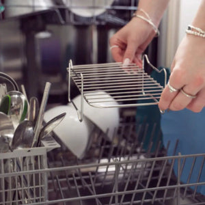 Woman loading stainless steel wire shelf insert into dishwasher basket, displaying Kitchen Academics air fryer accessory set with organized utensil compartments and multiple tiers