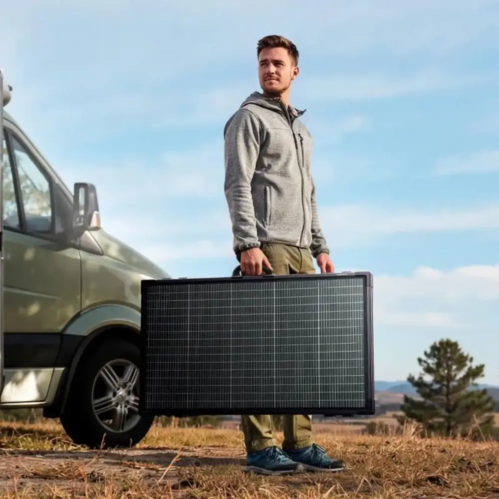 Man in grey hoodie and khaki pants holding large black bifacial solar panel in front of parked van in open landscape with clear blue sky