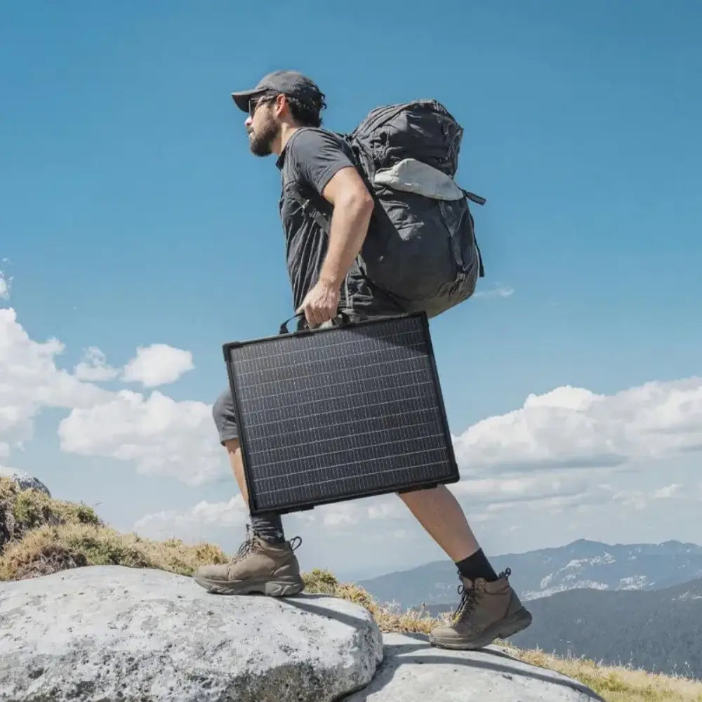 Man wearing gray shirt and cap standing on rocky mountain terrain holding black bifacial solar panel with backpack against blue sky with white clouds and forested mountains in background