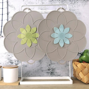 Beige silicone flower-shaped lid organizer with yellow and light blue decorative flower centers displayed on white stand next to white container and woven basket on concrete background