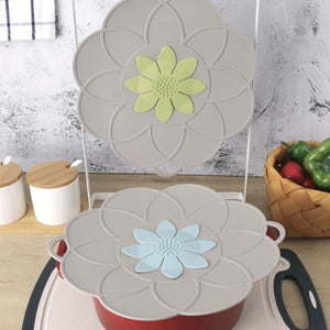 Flower-shaped silicone lids in cream with yellow and blue floral centers displayed on red and white cookware, shown in a kitchen setting with white containers and vegetable basket