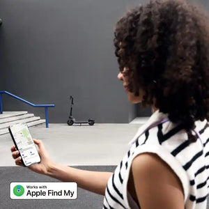 Woman with curly brown hair wearing white and black striped sleeveless top holding smartphone displaying Apple Find My app, looking at black Segway Ninebot electric scooter on white table with blue railing in background, demonstrating device tracking capability