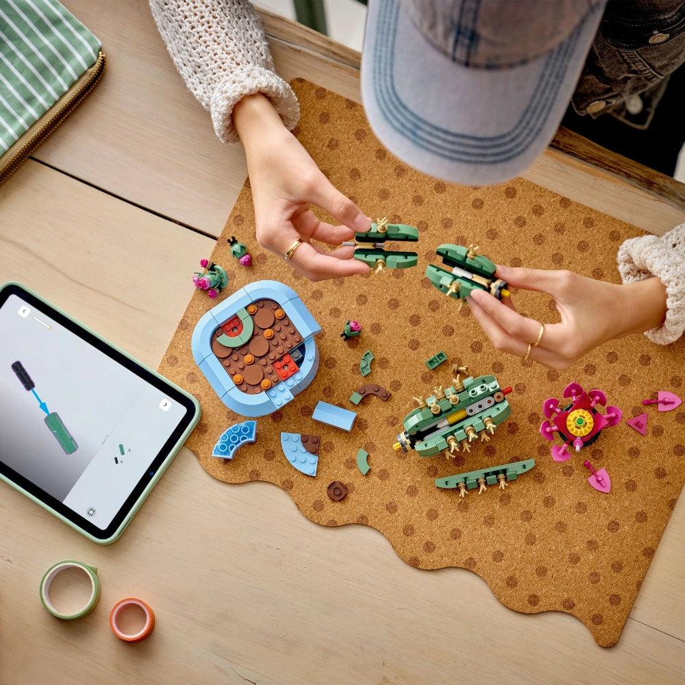 Child's hands assembling LEGO Flowering Cactus set with green segmented pieces and pink flower on cork mat, surrounded by colorful mini-figures and building elements, with tablet and craft supplies nearby on wooden surface