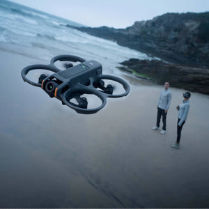 DJI Avata 2 FPV drone in dark gray with orange accents flying above a sandy beach near rocky cliffs with three people standing below operating the aircraft