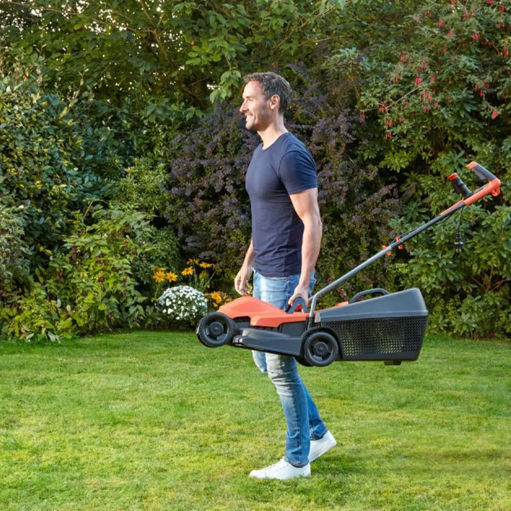 Man carrying a Black & Decker electric lawnmower with orange and black design in a garden with green lawn and flowering shrubs