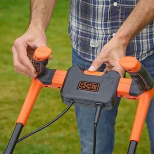 Man holding orange and black Black & Decker electric lawnmower with 32cm cutting width outdoors on grass, showing handle grips and power cable
