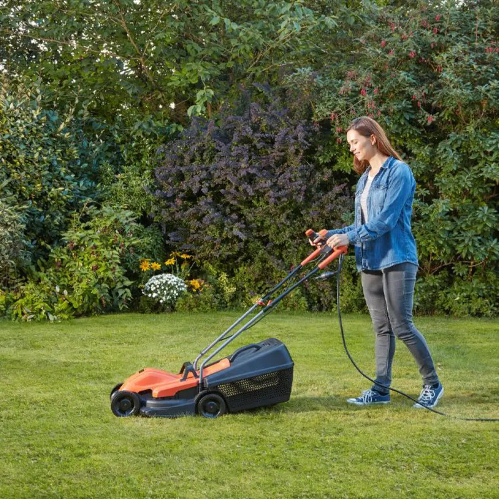 Woman operating a Black & Decker orange and black electric lawnmower on a green lawn with connected power cord, surrounded by flowering garden beds and green hedges