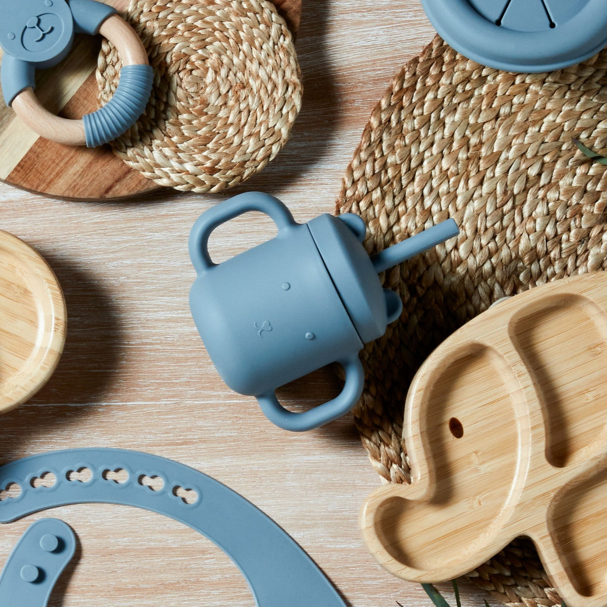 Flat lay product image of a blue-grey silicone sippy cup with handles surrounded by wooden bamboo plates, natural woven placemats, and blue silicone baby feeding accessories on a light wooden surface