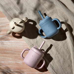 Three silicone sippy cups in cream, blue, and mauve pink arranged on a beige linen fabric with wooden surface, featuring handles, spouts, and embossed details