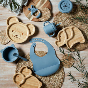 Bambino blue silicone sippy cup displayed among wooden bamboo plates shaped like bear and elephant heads, straw placemats, teething rings, and green foliage on light wood background