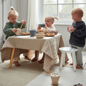 Three toddlers playing with wooden and silicone tableware around a draped table with natural light from large windows, featuring beige and neutral-toned play dishes and cups