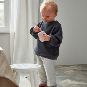 Toddler with blonde hair wearing dark blue knit sweater and cream pants holding pink silicone sippy cup with straw in bright room with white curtains and small white side table
