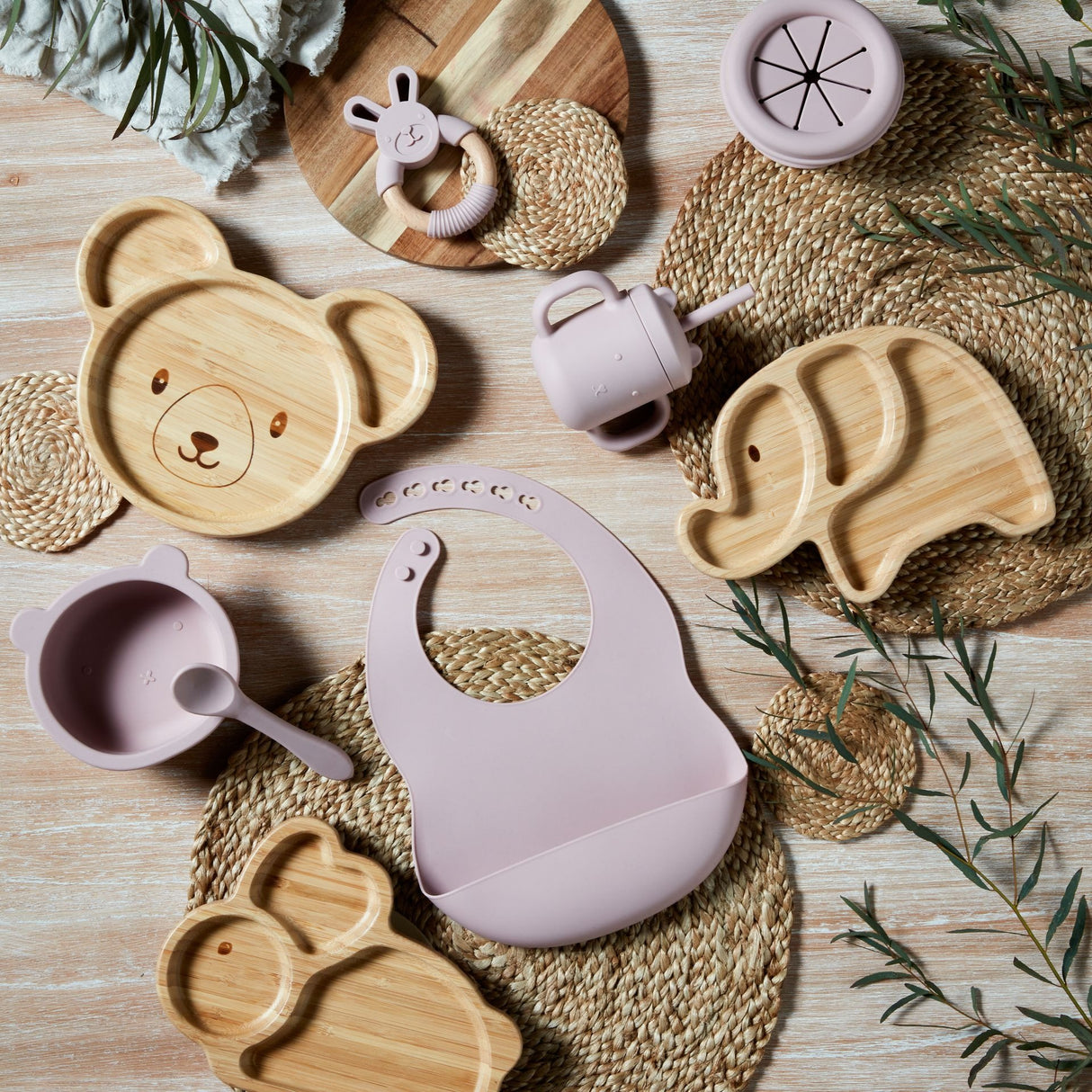 Overhead flat lay of Bambino Silicone Sippy Cup in soft pink surrounded by matching bamboo feeding plates with bear and elephant designs, purple silicone cup, bunny teether, woven placemats and green foliage on light wooden surface