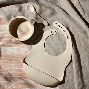 Bambino Natural silicone feeding set displayed on beige linen fabric featuring a bear-shaped bowl with lid, matching bib with suction cups, and spoon in cream white colour