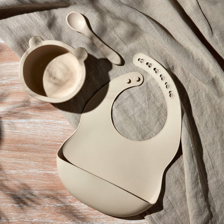 Bambino Natural silicone feeding set displayed on beige linen fabric featuring a bear-shaped bowl with lid, matching bib with suction cups, and spoon in cream white colour