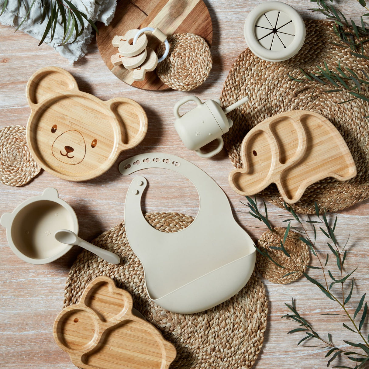Overhead flat lay of natural bamboo and silicone baby feeding set including bear-shaped wooden plates, elephant-shaped wooden plate, white silicone bib, cream ceramic cup, wooden spoon and accessories arranged on woven straw placemats with dried greenery on whitewashed wood surface