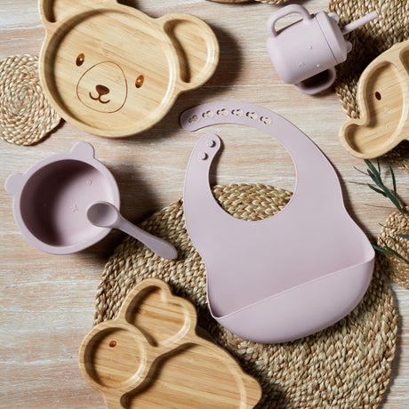 Bambino silicone feeding set in pink featuring a bib with snap closure, matching bowl with spoon, and wooden bamboo plates with bear design on woven placemats and light wood surface