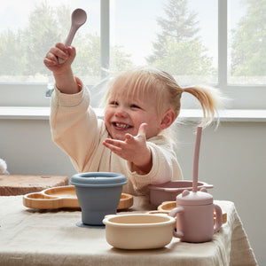 Young toddler with blonde hair eating from a silicone feeding set featuring a cream bowl, pink spoon, blue cup and pink sippy cup with handles on a wooden table by bright windows with forest view