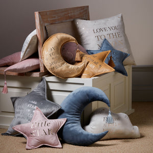 Bambino Velvet Star Cushion in pink with silver embroidered text displayed alongside coordinating nursery decor including moon and star-shaped pillows in blue, grey and cream velvet with tassels, styled in a wooden crate with white storage box