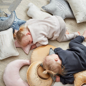Two sleeping babies on a beige textured rug surrounded by soft nursery products including cream and navy velvet star-shaped cushions, embroidered pillows, and knitted blankets in neutral tones