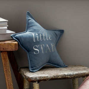 Blue velvet star-shaped cushion with "little STAR" text in white embroidery, displayed on a rustic wooden bench next to stacked books against a gray wall