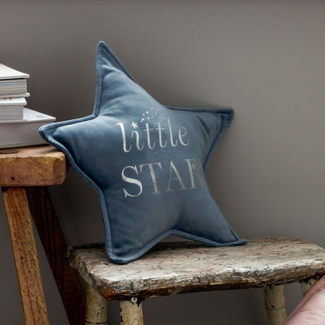 Blue velvet star-shaped cushion with "little STAR" text in white embroidery, displayed on a rustic wooden bench next to stacked books against a gray wall