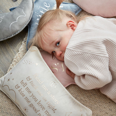Toddler cuddling a cream velvet rectangular cushion with rose gold text reading "Read me a story tell me you love me & miss me so good" surrounded by stars and moons, placed on a textured beige rug with gray and blue nursery items in background