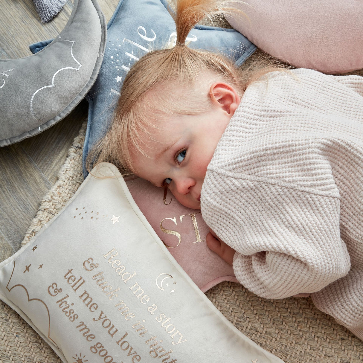 Toddler hugging cream velvet cushion with "Read me a story" text and star design, surrounded by coordinating nursery pillows in soft pastels on wooden floor