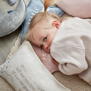Toddler hugging cream velvet cushion with "Read me a story" text and star design, surrounded by coordinating nursery pillows in soft pastels on wooden floor
